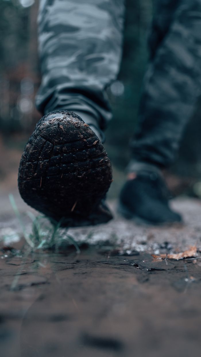hero-img-01 Close-up of a sneaker stepping on a wet, muddy path in a forest setting.